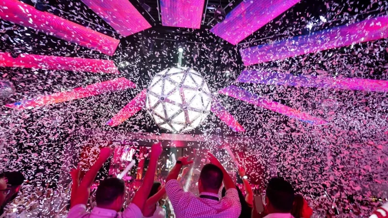 People dance at a party with purple lights, pink confetti, and a large disco ball overhead.