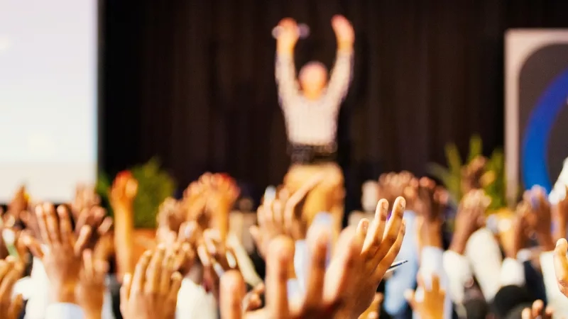 A person stands on stage as a crowd of cheering attendees raise their hands in excitement.