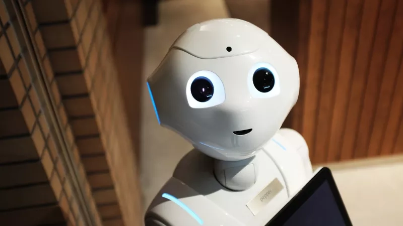 A white robot with blue eyes and a Pepper tag is holding a tablet against a brick wall.