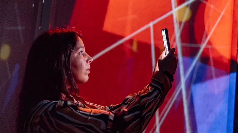Woman with phone in hand standing in front of red wall with abstract geometric lines projected on it.