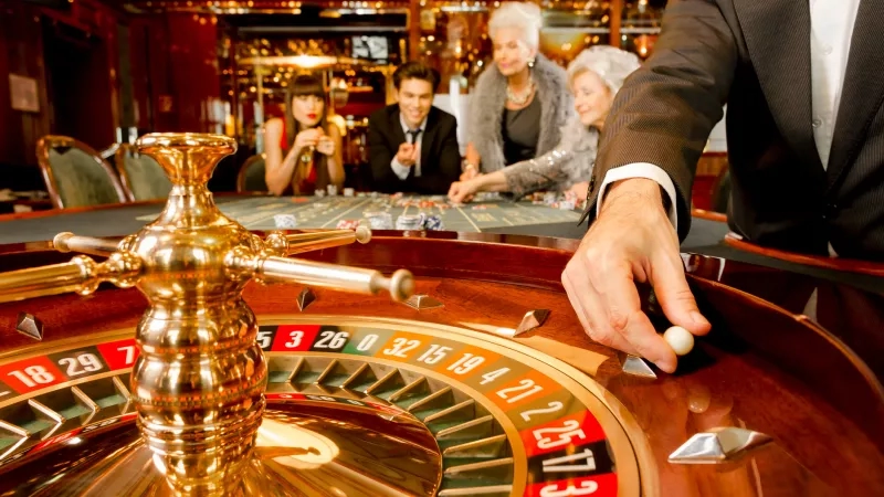 A dealer spins a roulette wheel in a casino while customers watch and wait for the ball to land.