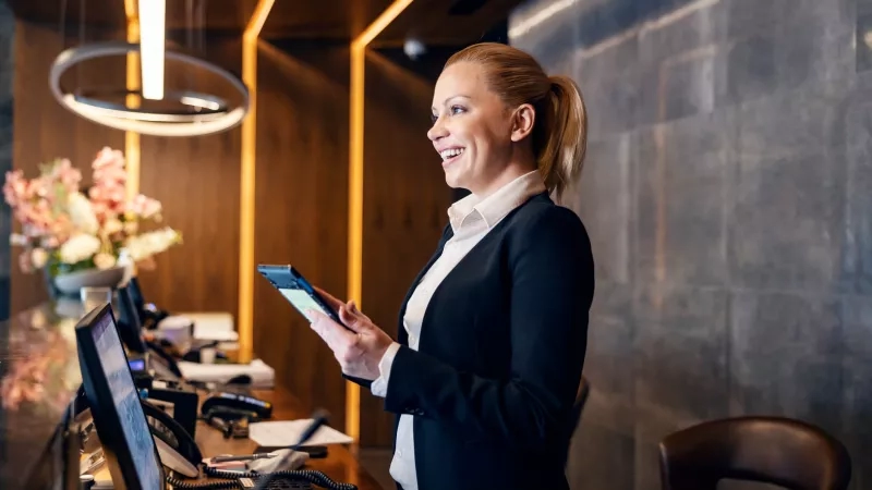 Hotel staff in lobby with tablet smiling at camera with flowers and desk in background.