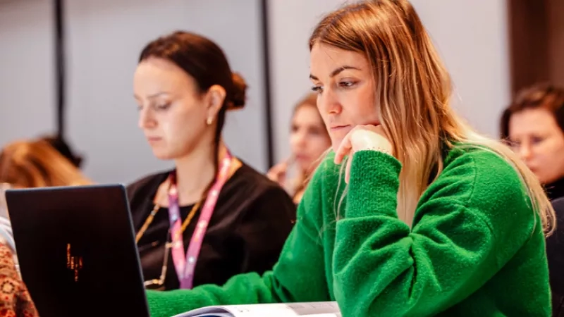 Two women sitting at a table with a laptop, one of them is wearing a green sweater.