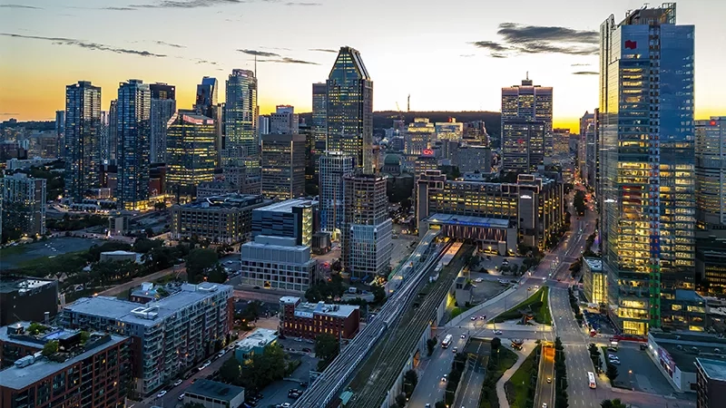 Aerial view of Montreal cityscape with tall buildings, a train track, and a busy road at sunset.