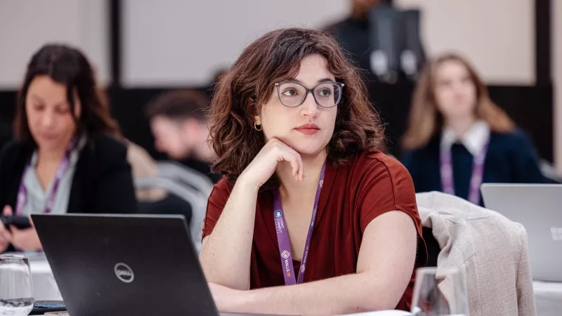 Woman wearing glasses and a purple lanyard sitting at a table with her laptop and a glass of water.