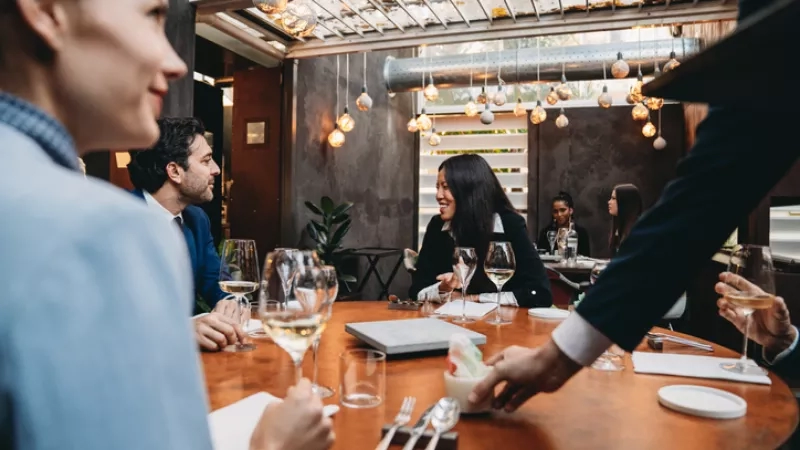 People in a restaurant with food, drinks, cutlery, and a table while a waiter is serving them.