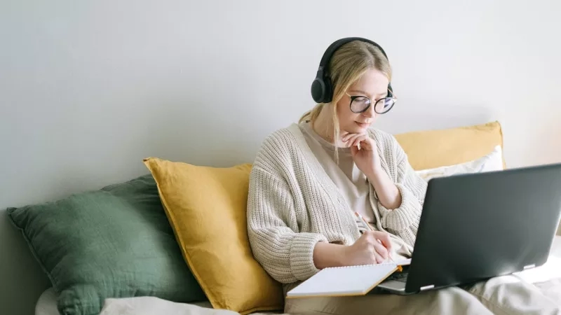 Woman sitting on bed with headphones, laptop, and notebook working on the side.