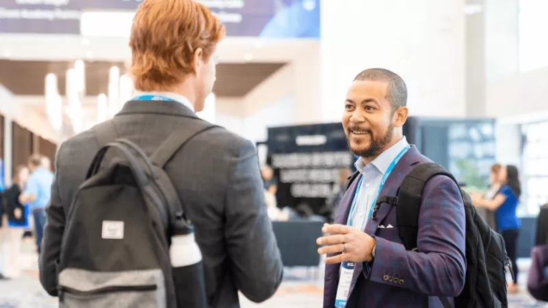 Two men talking in the hallway of a business travel conference with a banner in the background.
