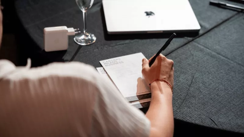 A person with a pen and notebook writing meeting minutes at a round table with a laptop.