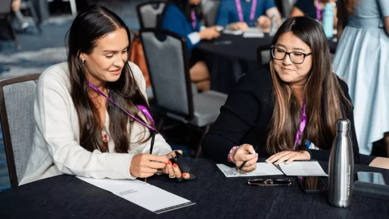 Two women sit at a table during a meeting, discussing and taking notes with a pen and paper.