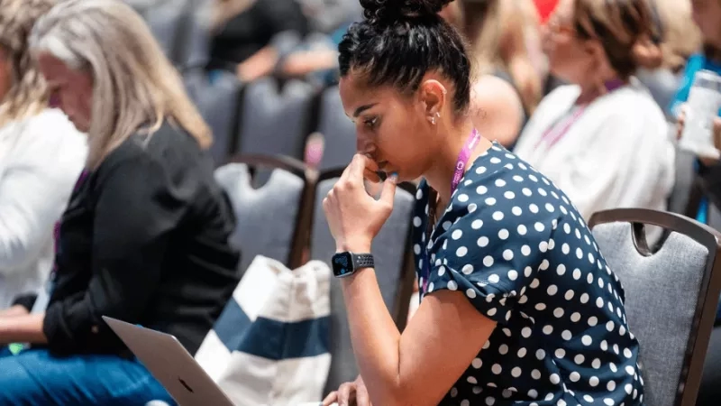 A woman working on her laptop at a conference with other women in the background.