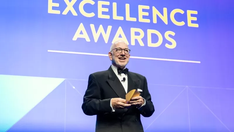 A man is standing on stage with a smile, holding an award in his hand, and wearing glasses.