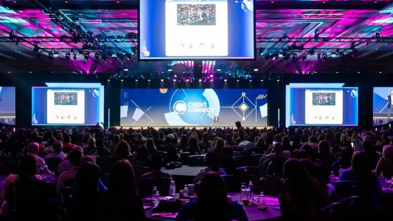 A large group of people are gathered in a room with purple lights and large screens on the wall.