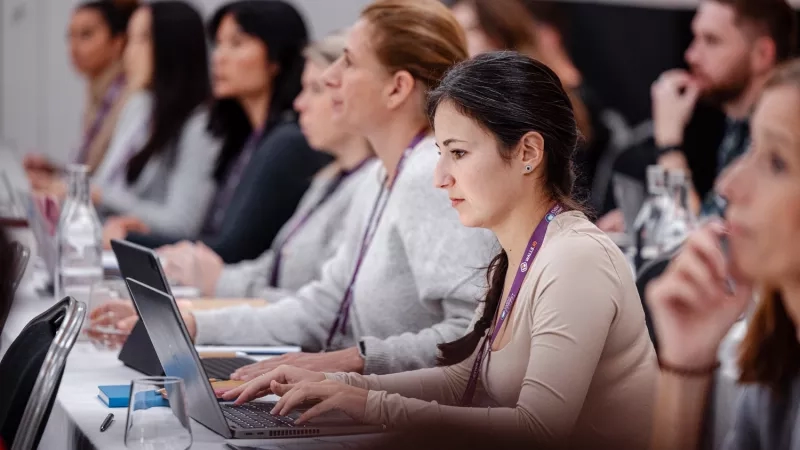 A group of women are sitting at a table and using laptops, with a man behind them.