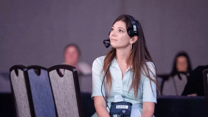 A woman with a headset and name tag, possibly an event planner, is seated at a conference.