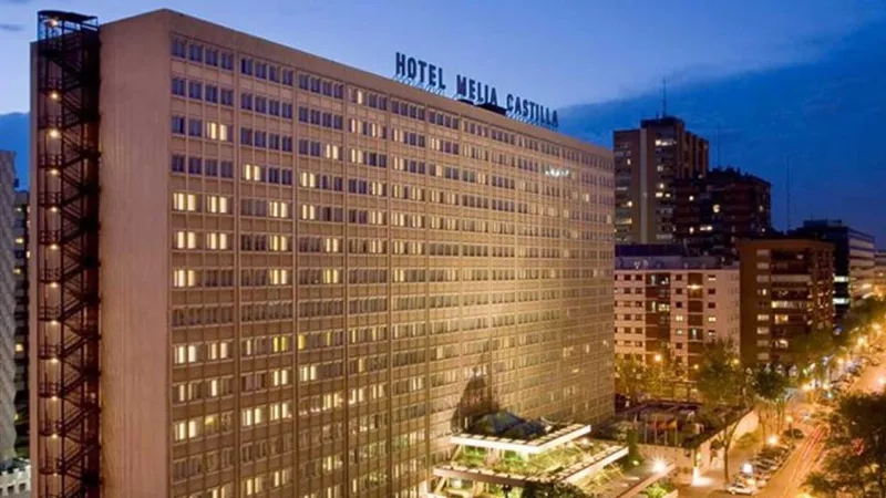 Hotel Melia Castilla, a tall building with many windows and a lit sign, stands in a cityscape at dusk.