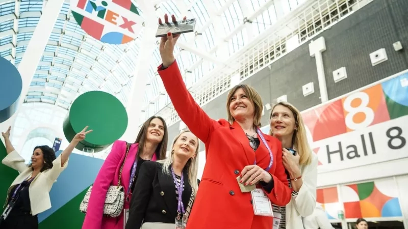 A group of women in formal attire take a selfie in the lobby of IMEX Frankfurt.