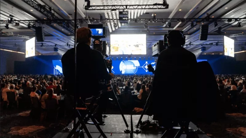 Two people recording an event with cameras and tripods, in front of a large crowd of people.