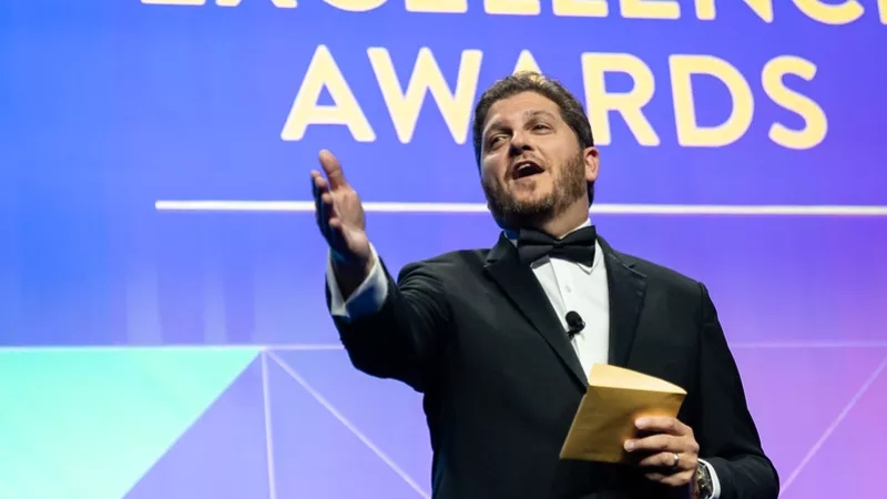 A man in formal attire is speaking while holding a piece of paper in front of a screen.