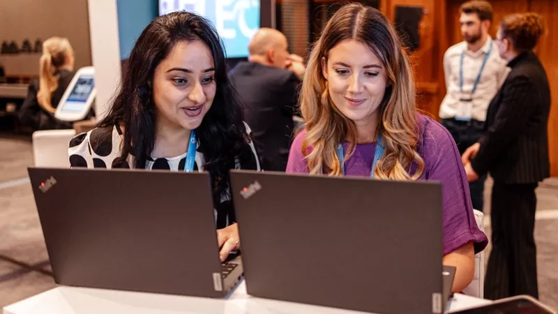 Two ladies working on laptops while others are standing and working in the background of an office.