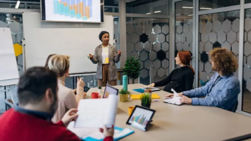 A group of people are sitting in a meeting with a woman giving a presentation on the screen.
