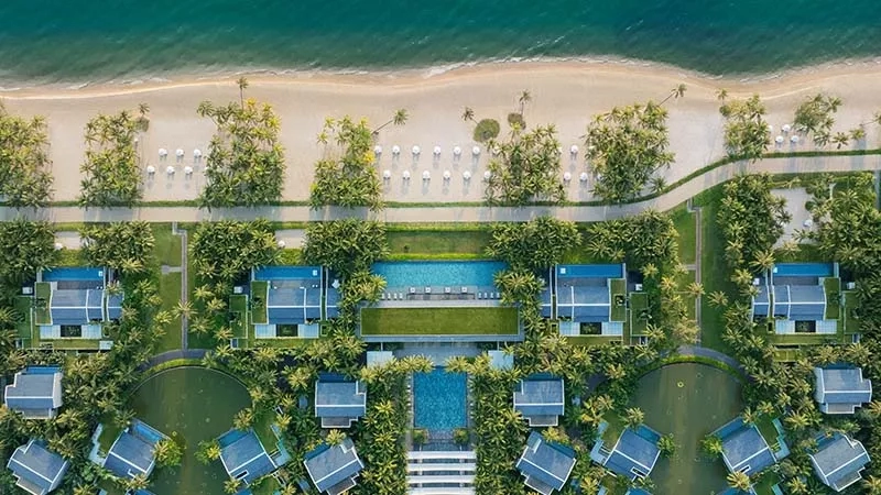 An aerial view of a resort featuring a pool, lush trees, and a sandy beach.