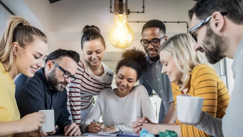 Group of colleagues gathered around a table with papers and a lightbulb hanging from the ceiling.