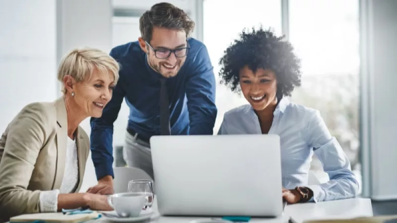 Three business people sitting in front of a laptop in a meeting at a desk in an office.