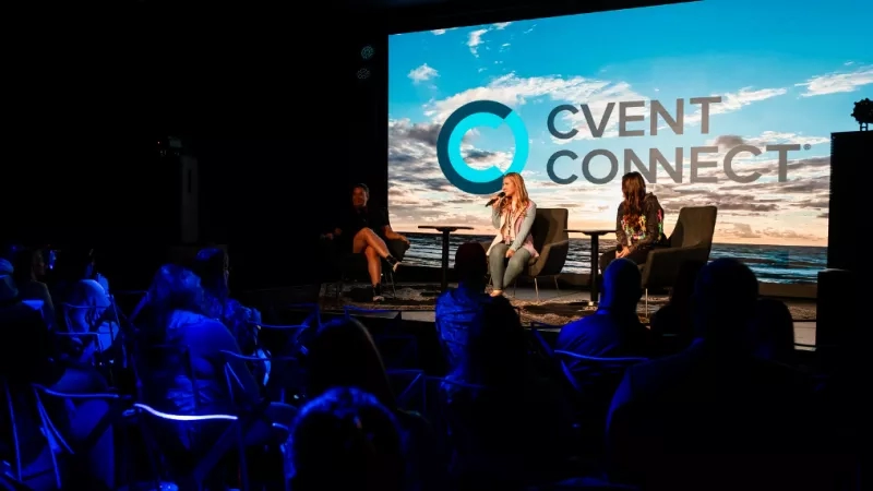 Three people sit on stage at an event with a crowd of people watching from chairs.