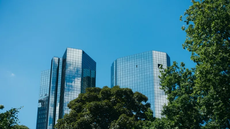 Twin Deutsche Bank towers in Frankfurt, Germany, surrounded by trees under a clear blue sky.