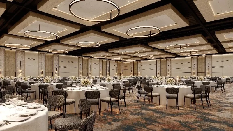 A large ballroom with round tables and chairs, illuminated by circular chandeliers and intricate ceiling design.