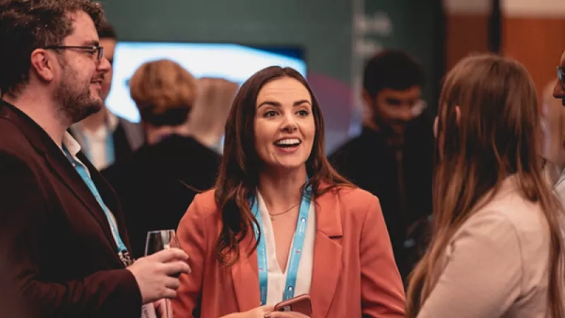A smiling woman holding a phone talking to two other people at a conference in a conference room.