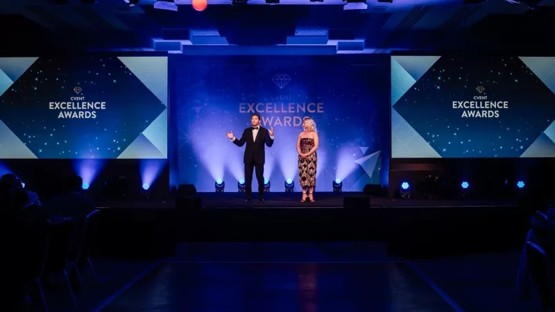 Man and woman standing on stage at Excellence Awards, with blue lighting and large screens displaying logos.