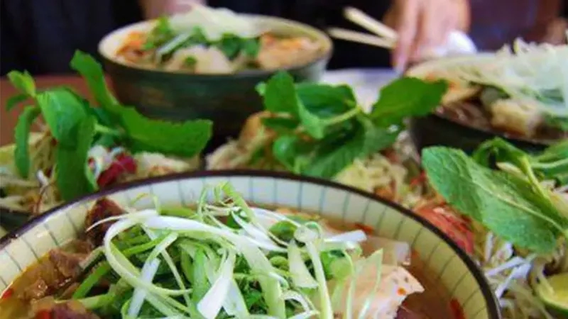 A bowl of noodles with green vegetables and herbs on a table with other bowls of food.