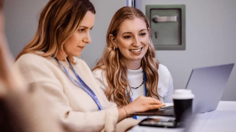 Two women are working together on a laptop, smiling, and enjoying a coffee.