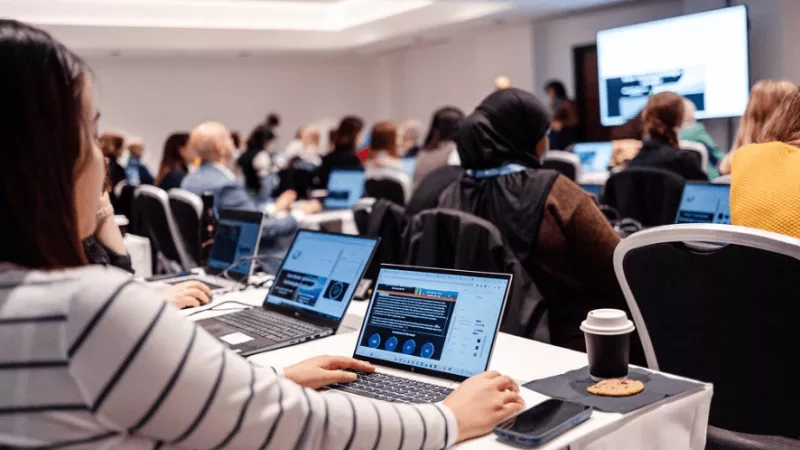 Woman sitting in front of laptop, surrounded by others, at conference in a room with large screen.