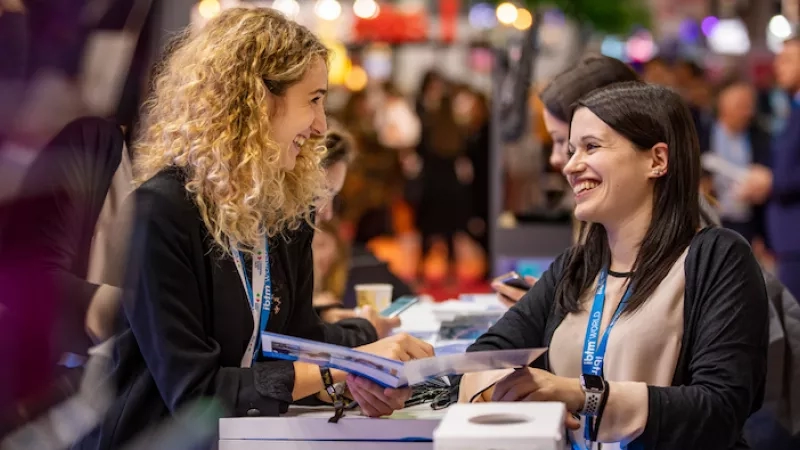 Two women chatting at a meeting with a blurred background of people and an event space.