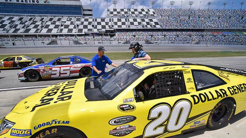 A racetrack with cars parked and people, one of which has the number 20 on it.