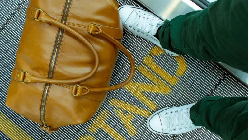 A person standing on an escalator with a tan leather travel bag on the ground.