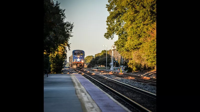 A blue train with a white star drives down tracks next to a walkway and lush green trees.