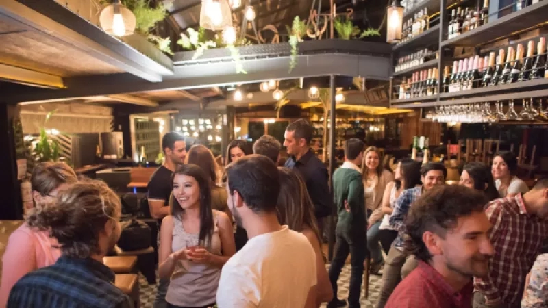 Group of people standing and talking in a bar with shelves full of bottles on the walls.