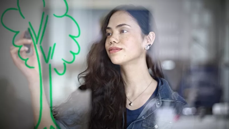 A woman in a denim jacket with long hair writes on a glass window with a marker.