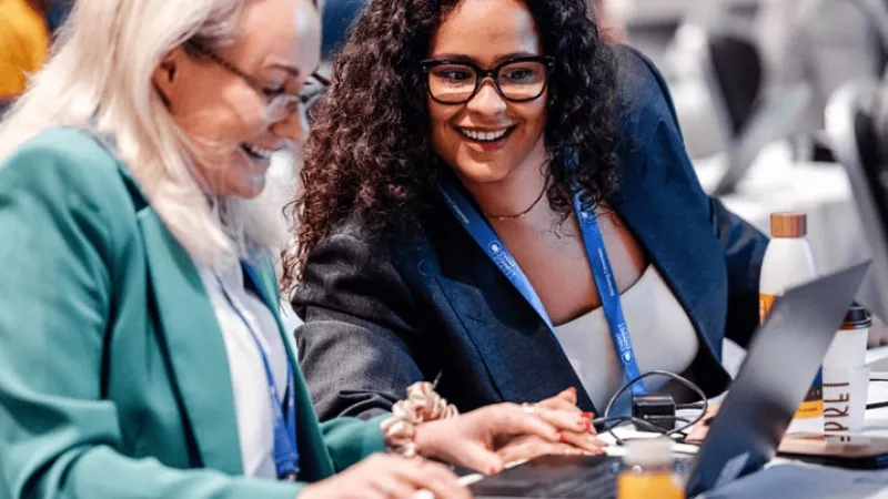 Two women wearing eyeglasses are sitting on chairs and working on laptops on a table in an office.