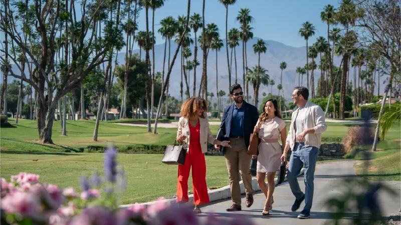 Four people walking on a path near flowers, palm trees, and a mountain in the background.