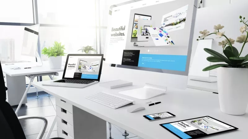 Office interior with monitor displaying website designs, and laptop on desk with a vase of flowers.