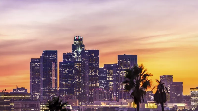 Cityscape of Los Angeles at sunset featuring tall buildings and palm trees against a colorful sky.