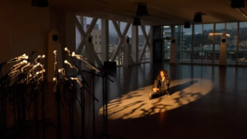 A person sits in a circle of light surrounded by many microphones in a large room.