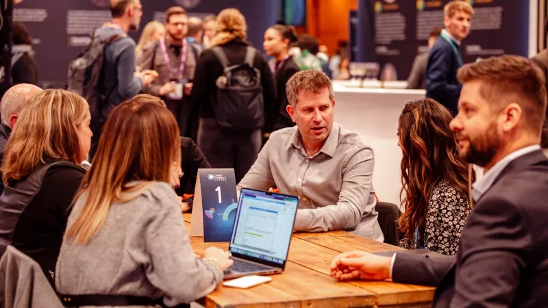 Several people sit at a table at a conference, with one person using a laptop.