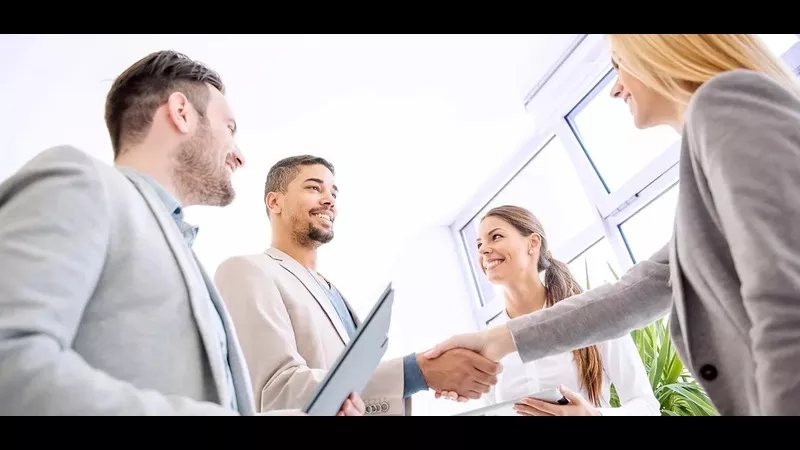 Group of business people shaking hands in an office with windows and plants in the background.
