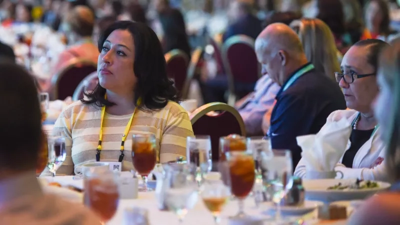 Woman with a yellow lanyard around her neck sits at a table with other people at a conference.